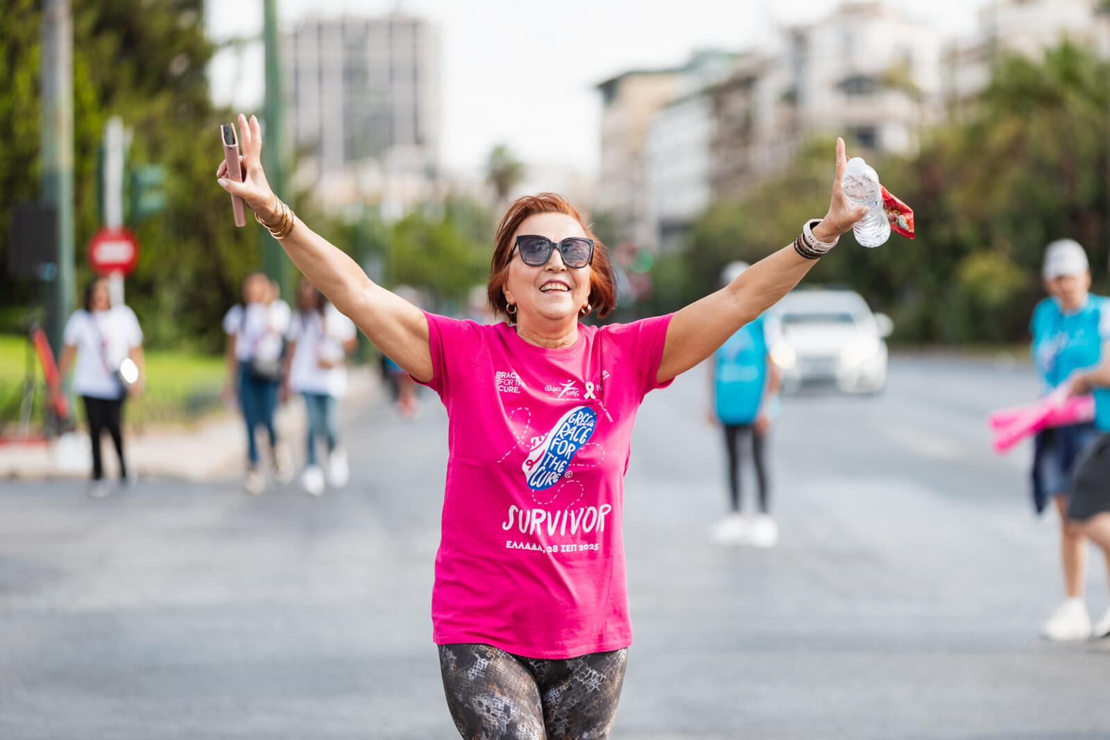 Γυναίκα survivor με ροζ μπλουζάκι χαμογελάει με τα χέρια απλωμένα στον περίπατο του Greece Race for the Cure®