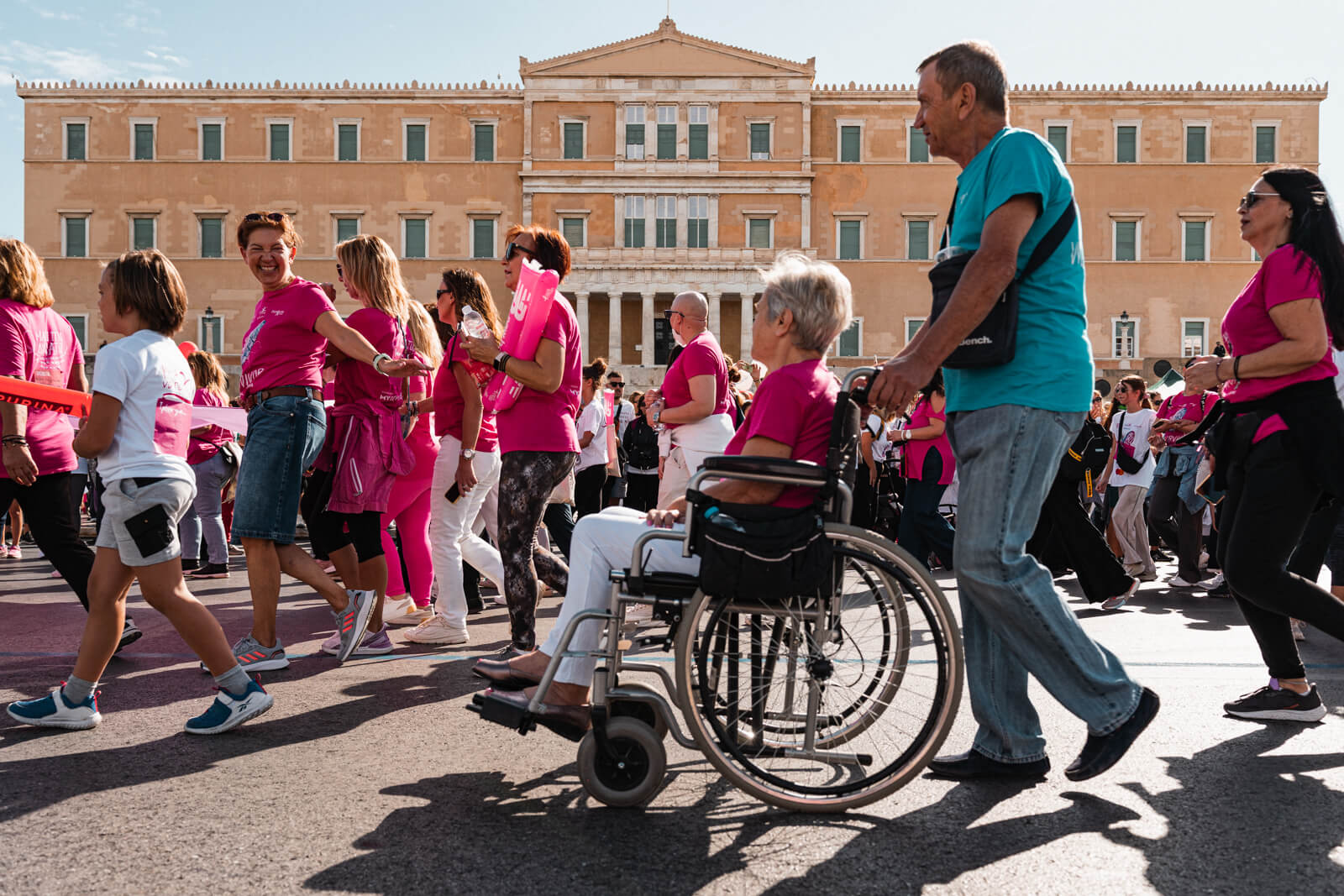 Στιγμές από τον περίπατο του Greece Race for the Cure®, γυναίκα με ροζ μπλουζάκι σε αμαξίδιο μαζί με άνδρα έξω από τη Βουλή